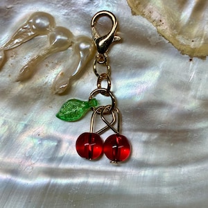 May include: A gold-toned metal lobster clasp charm featuring two translucent red spherical beads representing cherries, suspended from a gold ring alongside a small, textured, translucent green leaf-shaped charm made of acrylic or glass material.