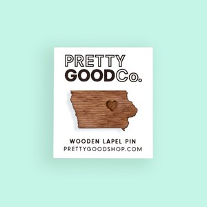May include: Wooden lapel pin shaped like the state of Iowa with a small heart cutout in the center. The pin is brown and has a wood grain texture. The text "PRETTY GOODCO." is printed in black at the top of the image. The text "WOODEN LAPEL PIN" is printed in black below the pin. The text "PRETTYGOODSHOP.COM" is printed in black below the text "WOODEN LAPEL PIN".
