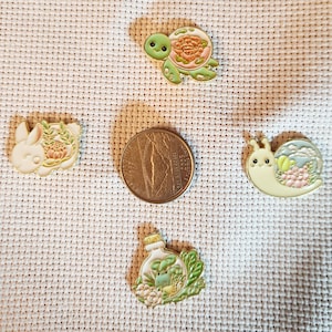 May include: An assortment of enamel pins featuring whimsical designs. The pins include a turtle with a floral shell, a bunny with flowers, a snail with a landscape, and a bottle with plants. A coin is included for size reference.