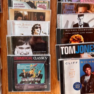 May include: A collection of music CDs in clear plastic jewel cases arranged in two columns. Visible titles include Commercial Classics, Tom Jones She's A Lady, Tom Jones Collection, Cliff Richard The Hit List, and Michael Crawford performs Andrew Lloyd Webber.