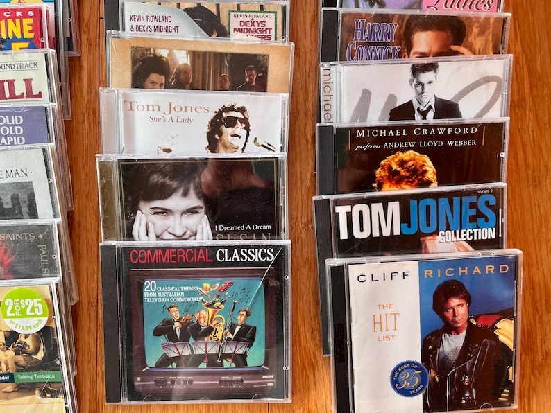 May include: A collection of music CDs in clear plastic jewel cases arranged in two columns. Visible titles include Commercial Classics, Tom Jones She's A Lady, Tom Jones Collection, Cliff Richard The Hit List, and Michael Crawford performs Andrew Lloyd Webber.