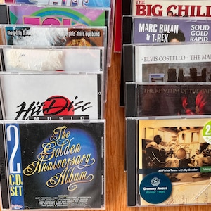 May include: A collection of music CDs in plastic jewel cases arranged in two columns. Visible titles include The Big Chill, Marc Bolan & T. Rex, Elvis Costello, The Rhythm of the Saints, Hit Disc Music, and The Golden Anniversary Album 2 CD set.