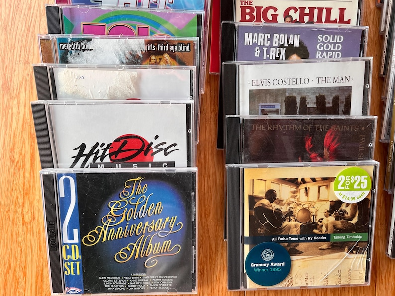 May include: A collection of music CDs in plastic jewel cases arranged in two columns. Visible titles include The Big Chill, Marc Bolan & T. Rex, Elvis Costello, The Rhythm of the Saints, Hit Disc Music, and The Golden Anniversary Album 2 CD set.