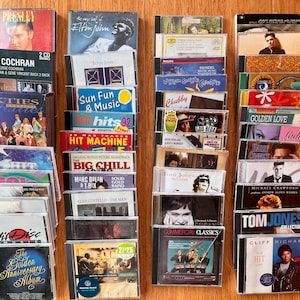 May include: A collection of music CDs arranged in four vertical columns. The cases feature various artists and titles including Elvis Presley, Elton John, Eddie Cochran, The Big Chill, Tom Jones, and Cliff Richard, with diverse graphic designs.