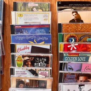May include: A collection of music CDs arranged in three vertical columns, featuring various album covers with artists like Lou Bega, Jimmy Barnes, and Mozart. The plastic jewel cases display diverse graphic designs, text, and photography.