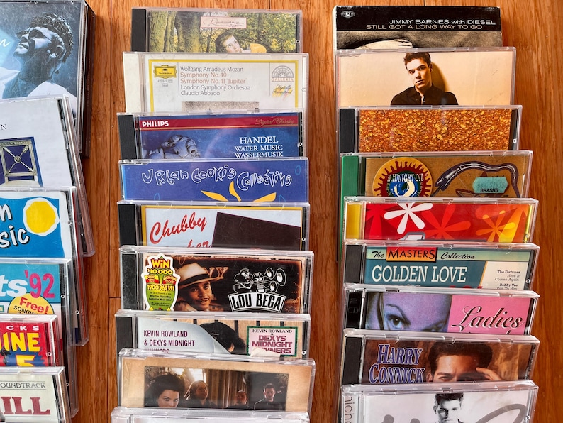 May include: A collection of music CDs arranged in three vertical columns, featuring various album covers with artists like Lou Bega, Jimmy Barnes, and Mozart. The plastic jewel cases display diverse graphic designs, text, and photography.