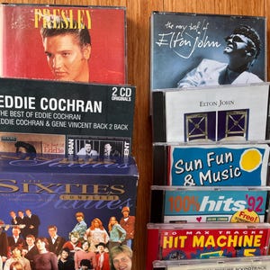 May include: A collection of music CDs and a box set titled The Sixties Complete. Visible titles include Elvis Presley, The Very Best of Elton John, Eddie Cochran, Sun Fun & Music, 100% Hits '92, and Hit Machine, featuring various artists and graphic designs.