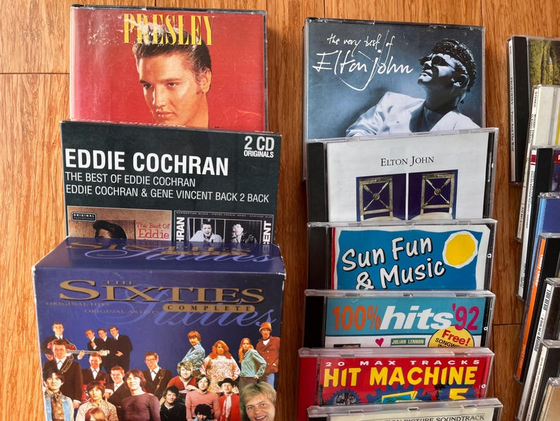 May include: A collection of music CDs and a box set titled The Sixties Complete. Visible titles include Elvis Presley, The Very Best of Elton John, Eddie Cochran, Sun Fun & Music, 100% Hits '92, and Hit Machine, featuring various artists and graphic designs.