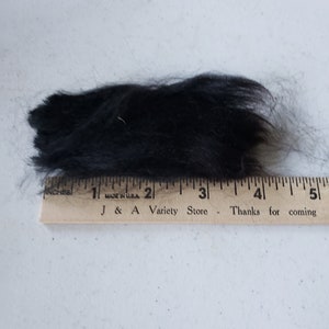 Op de afbeelding: Een close-up van een klomp zwart bont, gemeten met een houten liniaal met de tekst "J & A Variety Store - Thanks for coming".