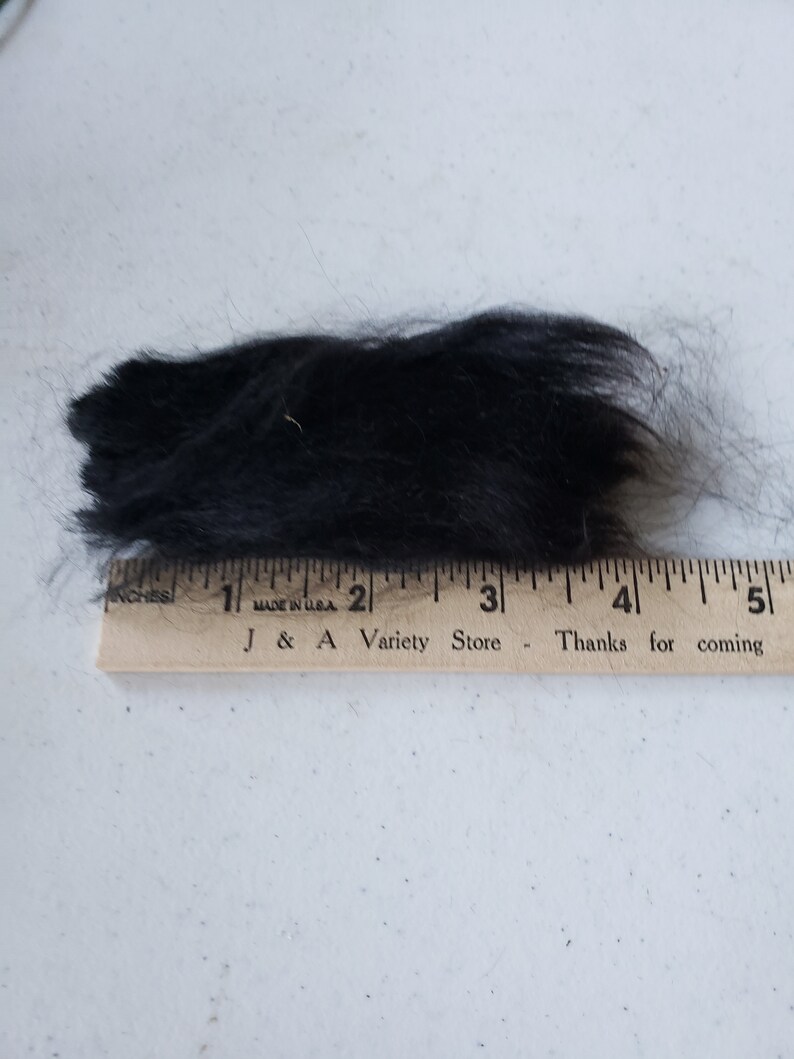 Op de afbeelding: Een close-up van een klomp zwart bont, gemeten met een houten liniaal met de tekst "J & A Variety Store - Thanks for coming".