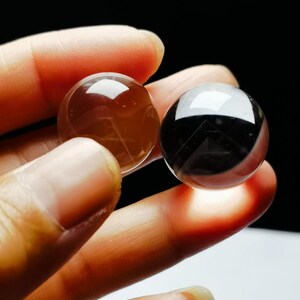 Op de afbeelding: Twee heldere, gepolijste kristallen bollen. Eén is een doorschijnende bruine kleur en de andere is zwart met een reflecterend oppervlak. De bollen worden tussen de vingers gehouden tegen een donkere achtergrond, wat hun gladde, afgeronde vormen benadrukt.