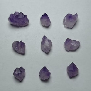 May include: Nine amethyst crystal clusters and points are arranged on a white surface. The crystals range in size and shape, with varying shades of purple and clear quartz.