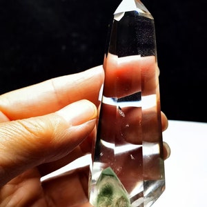 May include: A clear quartz crystal point with a green inclusion at the base. The crystal is faceted and has a pointed tip. It is held in a hand.