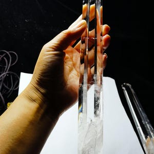 Może przedstawiać: Przezroczysty kryształ kwarcu, trzymany w dłoni, o fasetowanym, wydłużonym kształcie. Kryształ jest przezroczysty z wewnętrznymi wtrąceniami i spiczastym zakończeniem. Tło jest ciemne, podkreślając przejrzystość i kształt kryształu.