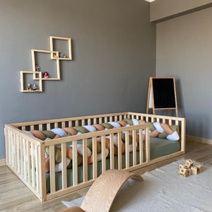 May include: A wooden playpen with a padded, braided bumper in shades of green, white, and orange. The playpen is set up in a room with a wooden floor and a gray wall with a set of wooden shelves.