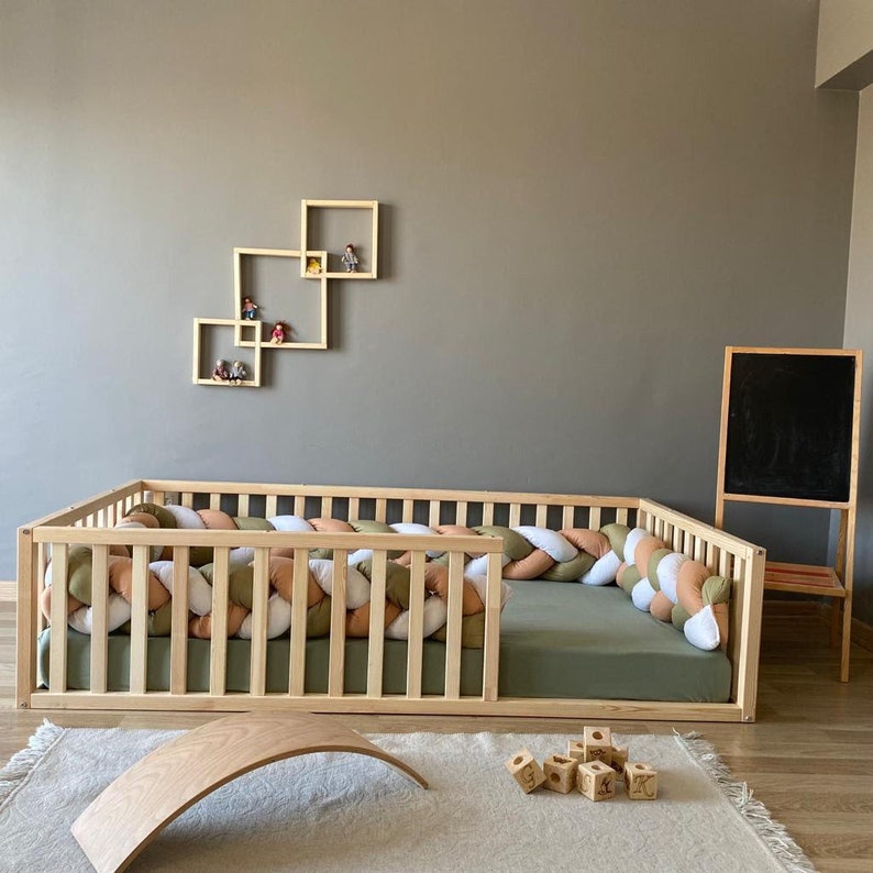 May include: A wooden toddler bed with a green mattress and a braided rope bumper in shades of white, beige, and green. The bed is surrounded by a wooden frame with slats. There are wooden blocks and a wooden balance board on the floor.