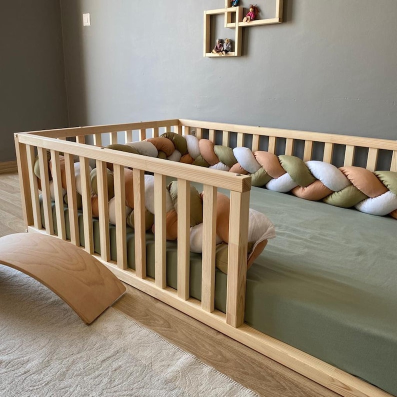 May include: A wooden toddler bed with a green mattress and a braided bumper made of white, beige, and green fabric. The bed is in a room with a light brown rug and a wooden wall shelf.