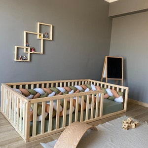 May include: A wooden crib with a braided rope bumper in shades of green, white, and orange. The crib has a slatted design and is surrounded by a wooden railing. The crib is set against a gray wall with a wooden shelf above it.