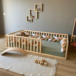 May include: A wooden toddler bed with a padded, braided bumper in shades of white, beige, and green. The bed has a green mattress and is surrounded by a wooden railing. There is a wooden rocking horse and a set of wooden blocks on the floor.
