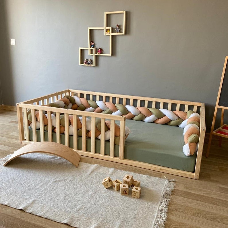 May include: A wooden toddler bed with a padded, braided bumper in shades of white, beige, and green. The bed has a green mattress and is surrounded by a wooden railing. There is a wooden rocking horse and a set of wooden blocks on the floor.
