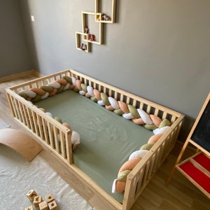 May include: A wooden crib with a green mattress and a white, beige, and green braided bumper. The bumper is made of soft fabric and is designed to protect the baby from bumps and falls.