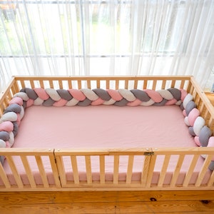 May include: A wooden baby playpen with a pink, white, and gray braided bumper pad. The playpen has a wooden frame and a pink sheet inside.