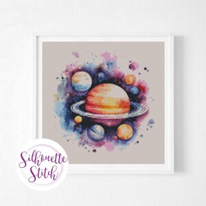 May include: A framed cross-stitch artwork depicting a planet with rings and other planets in a watercolour style. The colour scheme includes orange, blue, purple, and pink. The artwork is signed "Silhouette Stitch".