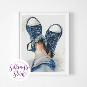 Puede incluir: Un par de zapatillas de lona azules con cordones blancos, gastadas y relajadas, con los dedos apuntando hacia arriba. Las zapatillas las lleva una persona que tiene las piernas cruzadas por los tobillos, y la persona lleva unos vaqueros azules.