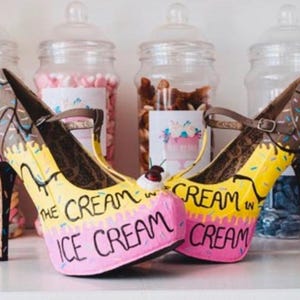 May include: Novelty high-heeled shoes with an ice cream theme. The shoes have a yellow base, pink accents, and brown ice cream details. The words "The Cream in Ice Cream" are written on the front. The heels are black.