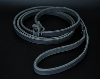 May include: A black leather camera strap with a metal buckle. The strap is coiled and laid flat on a black surface.