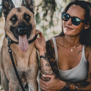 May include: A woman with tattoos on her arms is petting a German Shepherd dog. The dog is wearing a black collar and leash. The woman is wearing a white tank top, sunglasses, and a gold necklace with a circular pendant. The woman has a nose ring.