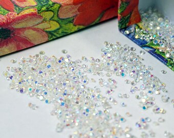May include: A small box with a floral pattern is open, revealing a pile of iridescent clear rhinestones. The box is made of cardboard and has a white interior.