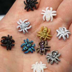 May include: Assortment of small, sparkly spider charms in various colors, including black, white, gold, silver, and blue. The charms are displayed on a hand.