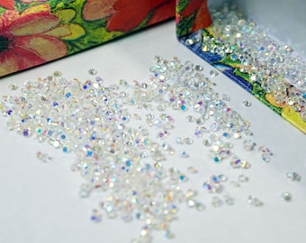 May include: A small box with a floral pattern is open, revealing a pile of iridescent clear rhinestones. The rhinestones are scattered on a white surface.