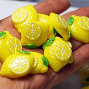 May include: A collection of yellow lemon-shaped resin cabochons with green leaves. The cabochons have a white raised design of a lemon slice.