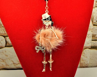 May include: A gold-tone necklace with a pendant featuring a cartoon woman wearing a hat, glasses, and a pink fur skirt. The necklace is on a black cord.