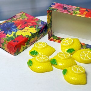 May include: A set of six yellow lemon-shaped soaps in a floral patterned box. The soaps are decorated with a white lemon slice and green leaves.