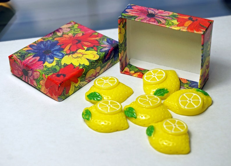 May include: A set of six yellow lemon-shaped soaps in a floral patterned box. The soaps are decorated with a white lemon slice and green leaves.