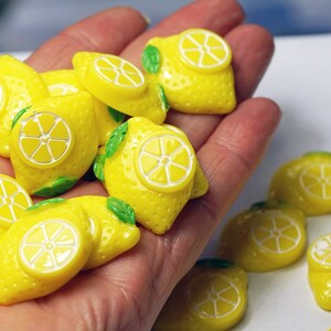 May include: A collection of yellow lemon-shaped resin cabochons with green leaves. The cabochons have a white center that resembles a lemon slice.