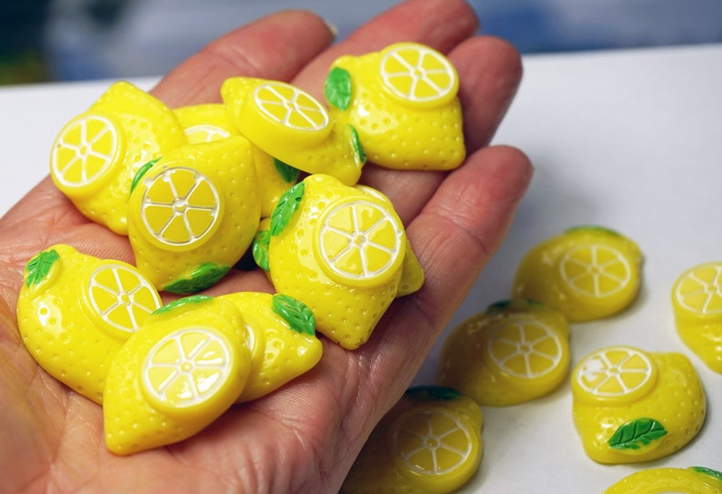 May include: A collection of yellow lemon-shaped resin cabochons with green leaves. The cabochons have a white center that resembles a lemon slice.