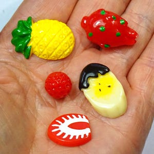 May include: A selection of fruit-shaped charms, featuring a yellow pineapple, a red strawberry, a banana with chocolate, a small red berry, and a red fruit slice with white detailing. These miniature fruit charms are likely used for crafting or jewellery making.