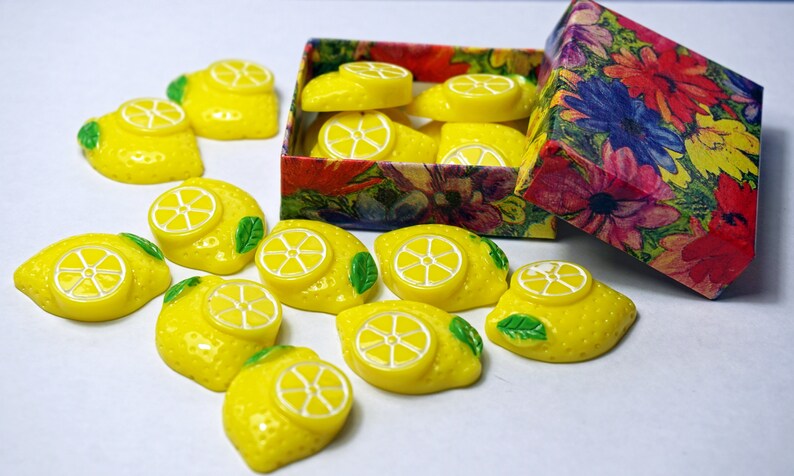 May include: A box of yellow lemon-shaped resin cabochons with green leaves. The box is decorated with a floral pattern.