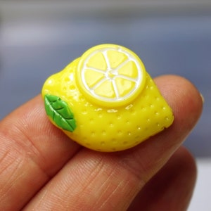 May include: A yellow lemon charm with a green leaf and a white lemon slice on top. The charm is made of polymer clay and is about 2 inches long.