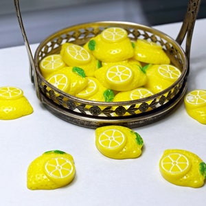 May include: A silver metal serving tray filled with yellow lemon-shaped candies. The tray is on a white surface with several more lemon candies scattered around it.