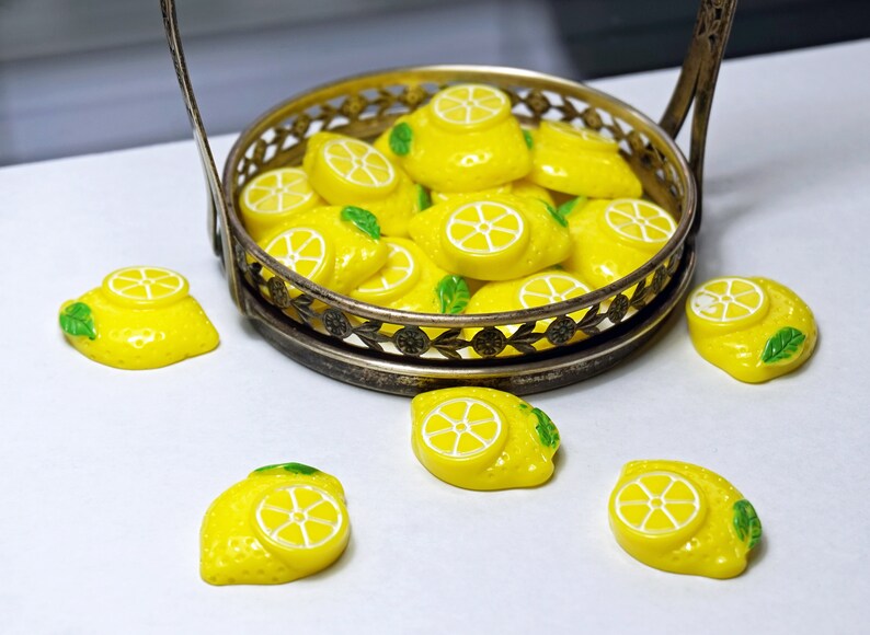 May include: A silver metal serving tray filled with yellow lemon-shaped candies. The tray is on a white surface with several more lemon candies scattered around it.