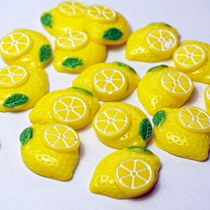 May include: A collection of 17 yellow lemon-shaped resin embellishments with green leaves. Each lemon has a white circle in the center, resembling a lemon slice.