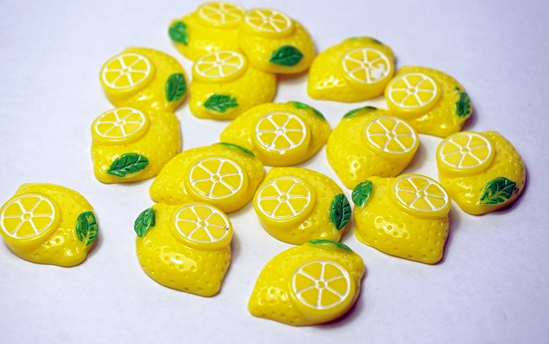May include: A collection of 17 yellow lemon-shaped resin embellishments with green leaves. Each lemon has a white circle in the center, resembling a lemon slice.
