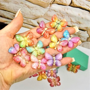 May include: A collection of colorful butterfly-shaped embellishments. The butterflies feature a glittery finish in various colors, including pink, orange, yellow, green, and blue. Each butterfly has gold-colored metal accents.