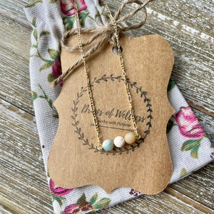 May include: A gold-colored necklace with three small, round, pastel-colored beads. The necklace is displayed on a brown tag with the text "Drops of Wellness" and a floral-patterned fabric pouch. The tag is tied with twine.
