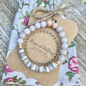 May include: A beaded bracelet with light pink and wooden beads, and shell-like accents, rests on a floral fabric pouch. A brown tag with the text "Props of Wellness Jewelry with Purpose" is tied with twine.